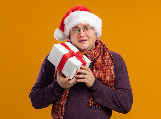 Blij volwassen man met bril en kerstmuts met cadeaupakket geïsoleerd op oranje muur package