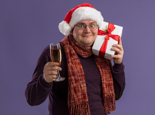 Blij volwassen man met bril en kerstmuts met sjaal om nek glas champagne te houden en hoofd aan te raken met cadeaupakket geïsoleerd op paarse muur