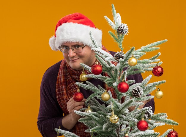 Blij volwassen man met bril en kerstmuts met sjaal om nek staan achter versierde kerstboom kijken camera geïsoleerd op een oranje achtergrond