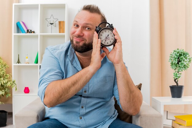 Blij volwassen Slavische man zit op fauteuil met wekker dicht bij het gezicht in de ontworpen woonkamer