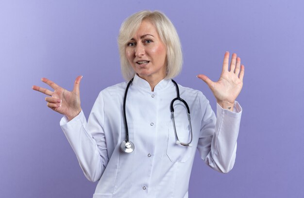 blij volwassen vrouwelijke arts in medische gewaad met stethoscoop gebaren acht met vingers geïsoleerd op paarse muur met kopie ruimte
