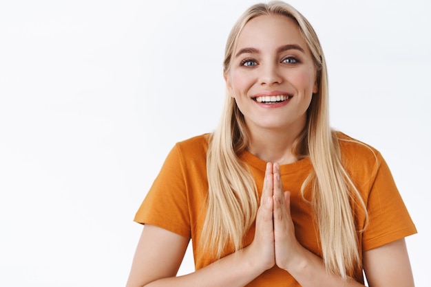 Blij vriendelijk vrolijk jong blond meisje draag oranje t-shirt druk de handpalmen tegen elkaar in bidden glimlachend bedankt voor hulp erg dankbaar dat je het op prijs stelt aanbod staande witte achtergrond opgetogen