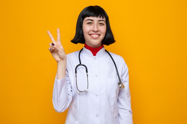 Blij, vrij Kaukasisch meisje in doktersuniform met stethoscoop gebaren overwinningsteken
