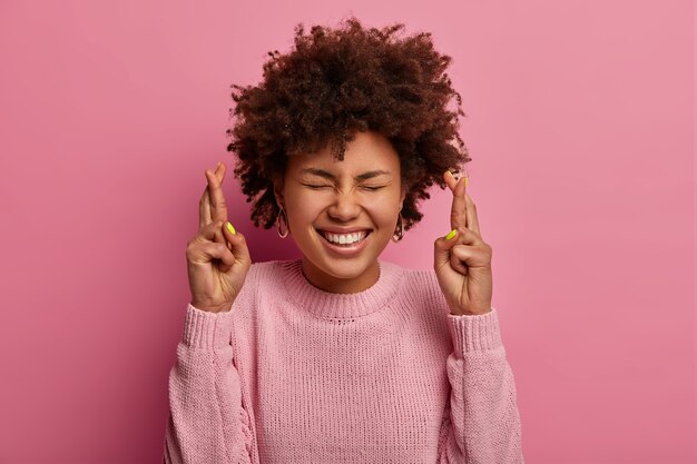 Blij vrouw met donkere huid staat met gekruiste vingers, anticipeert op belangrijk nieuws, gebaren binnen, heeft krullend haar, draagt een losse trui, maakt zoete begeerte, geïsoleerd over roze muur. Lichaamstaal