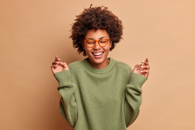 Gratis Foto blij vrouw steekt handen op heeft zorgeloze vrolijke uitdrukking sluit ogen glimlacht toothily draagt optische bril en trui geïsoleerd over beige muur
