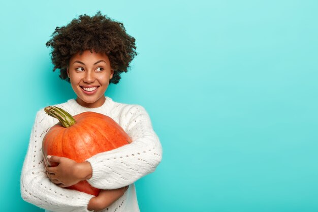 Blij vrouwtje heeft afro-kapsel, houdt grote pompoen vast, gebruikt gezond product voor het bereiden van biologische maaltijden, kijkt vrolijk weg, gekleed in trui