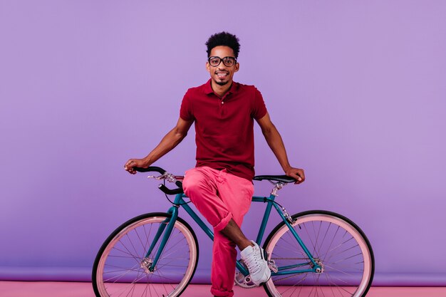 Blij zwart mannelijk model in roze broek zittend op de fiets. Binnen schot van lachende Afrikaanse jongen in geïsoleerde glazen.