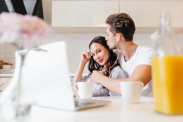 Blij zwartharig meisje genieten van favoriete muziek met vriendje met behulp van laptop. Indoor portret van schattige jonge vrouw rusten met echtgenoot met glas sap op de voorgrond.