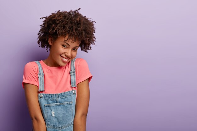 Blije Afro-Amerikaanse vrouw heeft een vrolijke verlegen gezichtsuitdrukking, drukt positieve emoties uit, draagt een roze t-shirt en denim sarafan, modellen over paarse muur, kopie ruimte opzij