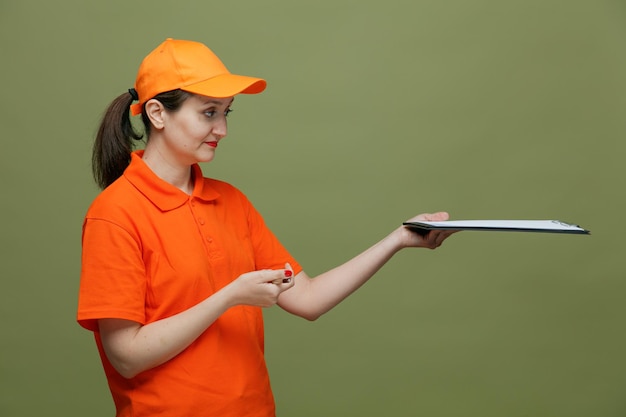 Blije bezorger van middelbare leeftijd, gekleed in uniform en pet in profielweergave met pen uitrekkend klembord uit kijkend naar kant geïsoleerd op olijfgroene achtergrond