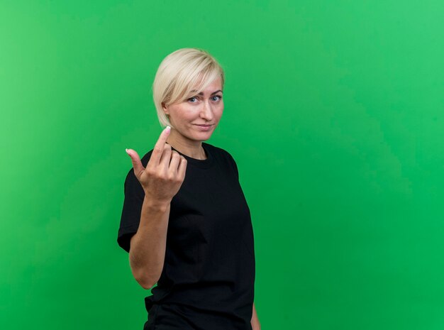Blije blonde Slavische vrouw van middelbare leeftijd die zich in profielmening bevindt die kom hier gebaar doet dat op groene muur met exemplaarruimte wordt geïsoleerd