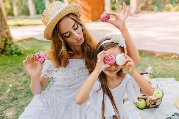 Blije blonde vrouw met lichte make-up die met liefde naar dochter kijkt die met koekjes speelt. Buitenportret van een klein meisje met snoepjes zoals een bril in de buurt van een mand met groene appels.