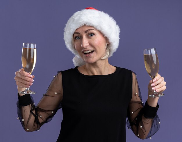 Blije blonde vrouw van middelbare leeftijd die Kerstmishoed draagt die camera bekijkt die twee glazen champagne houdt die op purpere achtergrond wordt geïsoleerd