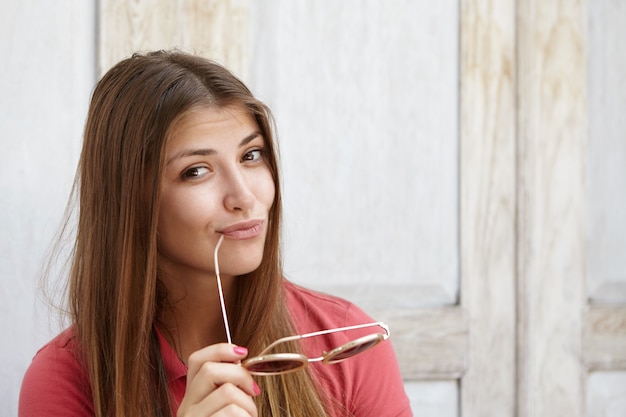 Blije en vrolijke jonge blanke vrouw met lang haar dat haar lippen pruilt, tinten vasthoudt en lippen aanraakt met de punt van de tempel, met flirten en mysterieuze blik, poseren geïsoleerd op houten muur