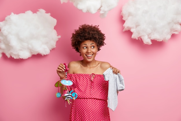 Blije etnische vrouw poseert met romper en mobiel, wordt binnenkort moeder, kijkt gelukkig opzij, heeft een dikke buik, draagt een polka dot-jurk, geïsoleerd op een roze muur, witte wolken boven het hoofd