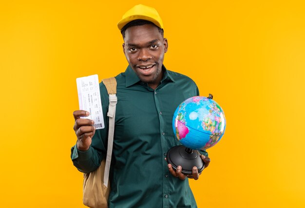 Blije jonge Afro-Amerikaanse student met pet en rugzak houdt vliegticket en globe geïsoleerd op oranje muur met kopieerruimte