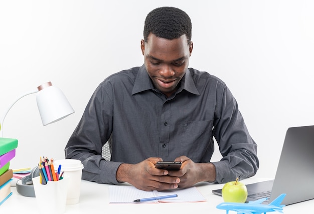 Blije jonge afro-amerikaanse student zit aan een bureau met schoolhulpmiddelen die de telefoon vasthouden en kijken