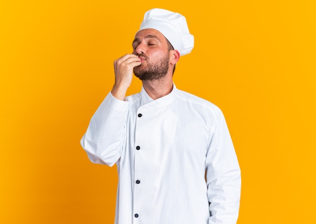 Blije jonge blanke mannelijke kok in chef-kokuniform en pet doet heerlijk gebaar met gesloten ogen