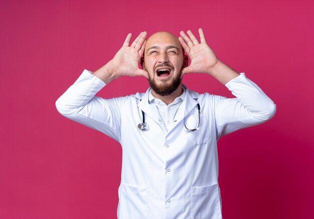 Blije jonge kale mannelijke arts die medische mantel en stethoscoop draagt die het gebaar van konijntjesoren tonen dat op roze muur wordt geïsoleerd