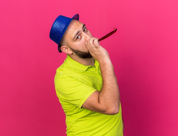 Blije jonge man met een blauwe feestmuts die een feestfluitje blaast dat op een roze muur wordt geïsoleerd