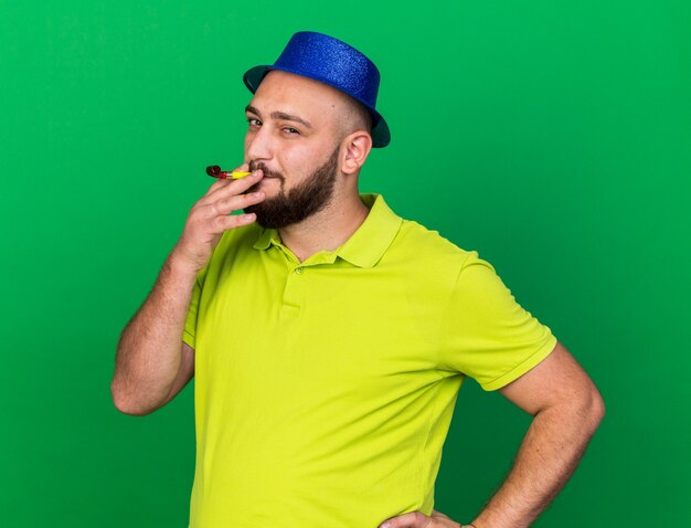 Blije jonge man met een blauwe feestmuts die een feestfluitje blaast en hand op de heup zet geïsoleerd op een groene muur
