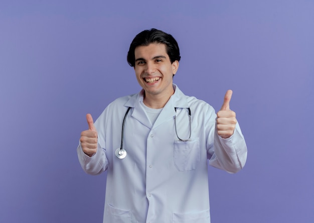 Blije jonge mannelijke arts die medische mantel en stethoscoop draagt die duimen toont die omhoog op purpere muur met exemplaarruimte worden geïsoleerd