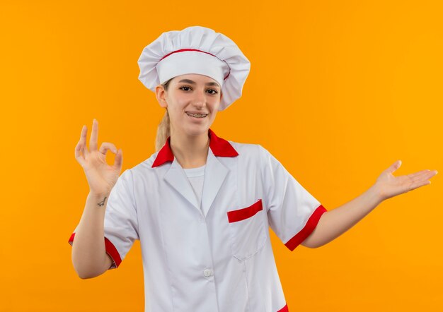 Blije jonge mooie kok in eenvormige chef-kok met tandsteunen die ok teken doen en lege die hand op oranje ruimte tonen tonen