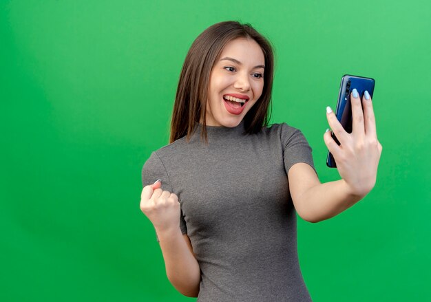Blije jonge mooie vrouw die en mobiele telefoon houdt en balde vuist bekijkt die op groene achtergrond met exemplaarruimte wordt geïsoleerd