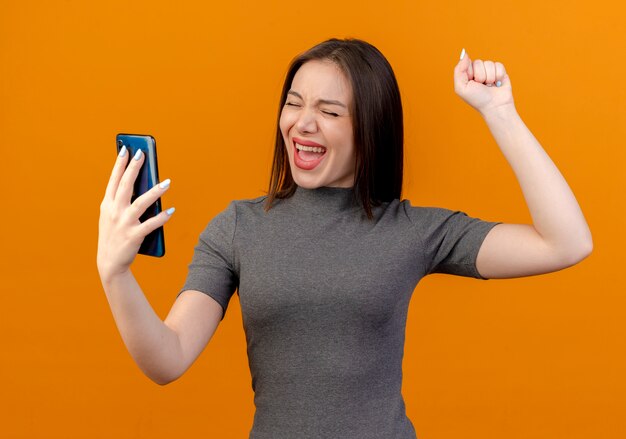 Blije jonge mooie vrouw die mobiele telefoon houdt en vuist met gesloten ogen opheft die op oranje achtergrond worden geïsoleerd