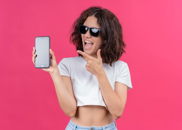 Blije jonge mooie vrouw die zonnebril draagt en mobiele telefoon houdt die op geïsoleerde roze muur met exemplaarruimte richt