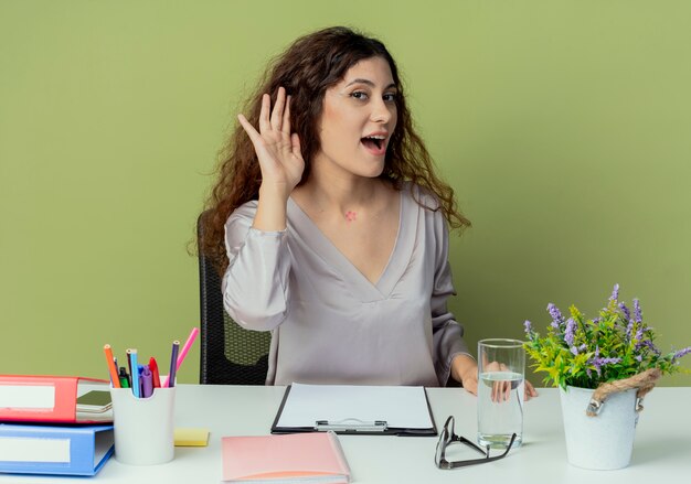 Blije jonge mooie vrouwelijke beambtezitting bij bureau met bureaushulpmiddelen die luister grsture tonen die op olijfachtergrond wordt geïsoleerd