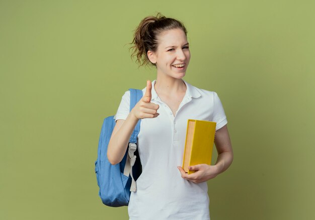 Blije jonge mooie vrouwelijke student die het boek van de achterzakholding draagt en op camera richt die op groene achtergrond met exemplaarruimte wordt geïsoleerd