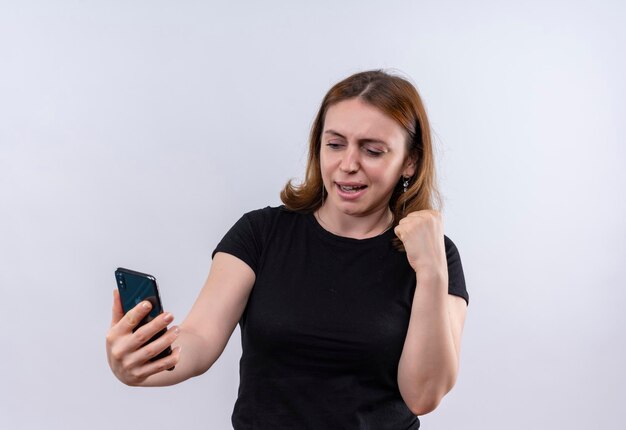 Blije jonge toevallige vrouw die mobiele telefoon houdt en vuist opheft op geïsoleerde witte ruimte met exemplaarruimte