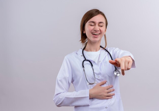 Blije jonge vrouwelijke arts die medische mantel en stethoscoop draagt en met gesloten ogen met exemplaarruimte richt