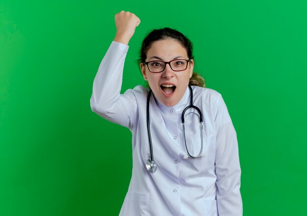 Blije jonge vrouwelijke arts die medische mantel en stethoscoop en bril draagt die ja gebaar doet dat op groene muur met exemplaarruimte wordt geïsoleerd