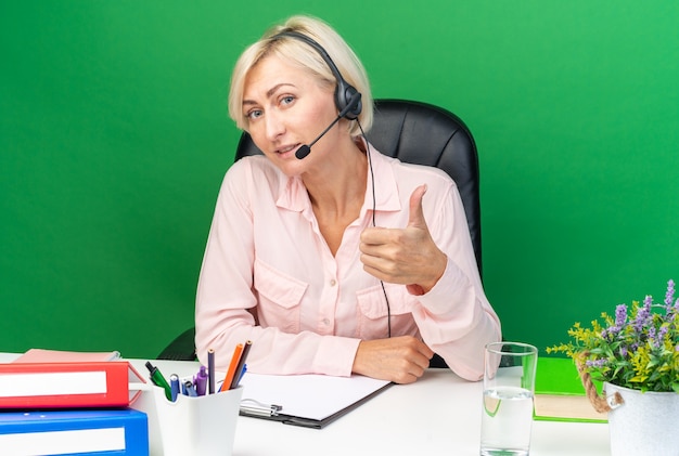 Blije jonge vrouwelijke callcentermedewerker met een headset die aan tafel zit met kantoorhulpmiddelen die duim omhoog laten zien