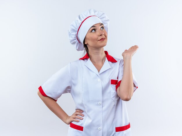 Blije jonge vrouwelijke kok die chef-kok uniform draagt die hand uitspreidt die op witte achtergrond wordt geïsoleerd