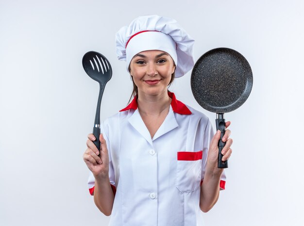 Blije jonge vrouwelijke kok die een chef-kok uniform draagt en een spatel vasthoudt met een koekenpan geïsoleerd op een witte achtergrond