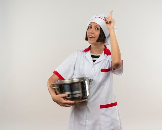 Blije jonge vrouwelijke kok in de pot die van de chef-kok eenvormige holding vinger opheft die op wit wordt geïsoleerd