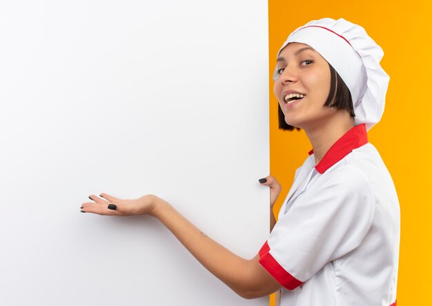 Blije jonge vrouwelijke kok in eenvormige chef-kok die zich dichtbij witte muur bevindt en met hand wijst naar het geïsoleerd op oranje