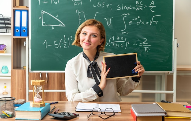 Blije jonge vrouwelijke leraar zit aan tafel met schoolgereedschap met mini-bord in de klas