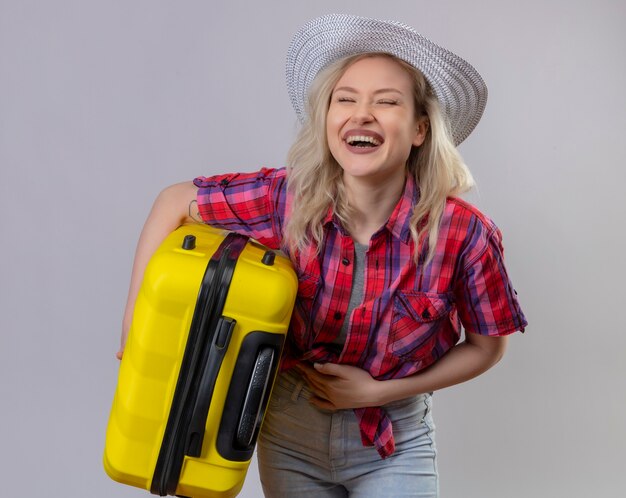 Blije jonge vrouwelijke reiziger die rood overhemd in de koffer van de hoedenholding draagt, legt hand op buik op geïsoleerde witte muur