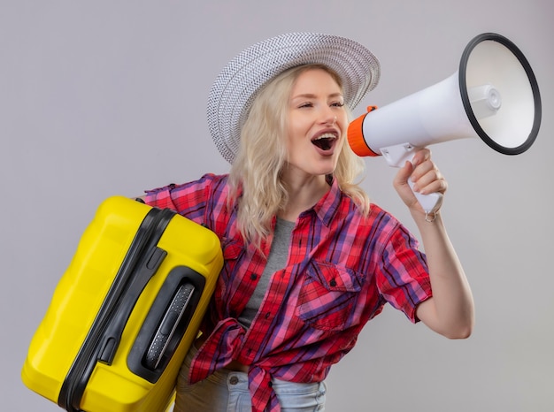 Blije jonge vrouwelijke reiziger die rood overhemd in de koffer van de hoedenholding draagt, spreekt door luidsprekers op geïsoleerde witte muur