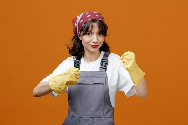 Blije jonge vrouwelijke schoonmaker met uniforme rubberen handschoenen en bandana die naar de camera kijkt en een boksgebaar toont dat op een oranje achtergrond wordt geïsoleerd