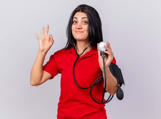 Blije jonge zieke vrouw die een stethoscoop draagt en kijkt naar de voorkant die haar druk meet met een bloeddrukmeter die ok teken doet dat op witte muur wordt geïsoleerd