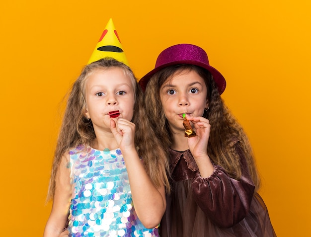 Blije kleine mooie meisjes met feestmutsen die feestfluitjes blazen geïsoleerd op een oranje muur met kopieerruimte