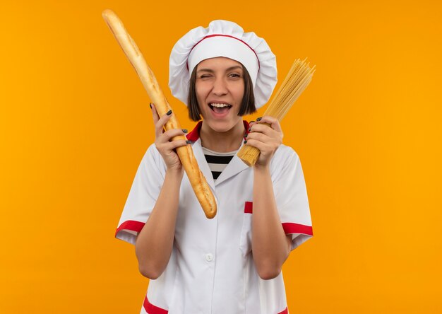 Blije knipogende jonge vrouwelijke kok in het broodstokje van de chef-kok eenvormige holding en spaghettideegwaren die op sinaasappel worden geïsoleerd