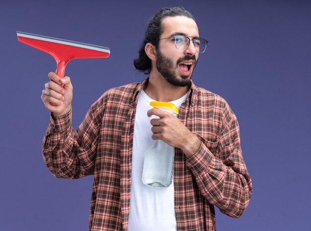 Gratis foto blije knipperde jonge knappe schoonmaakster die t-shirt draagt die zwabberhoofd met sproeifles houdt die op blauwe muur wordt geïsoleerd