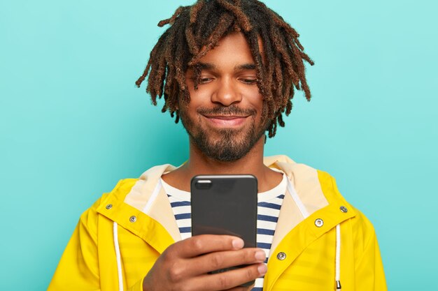 Blije man met donkere huid, heeft dreadlocks, kijkt positief naar smartphone, bekijkt foto's, draagt gele regenjas