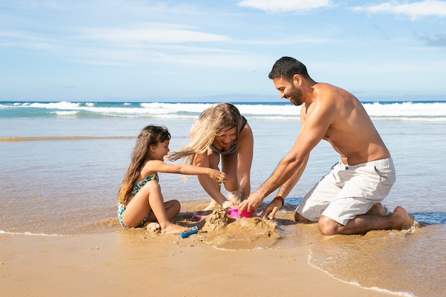 Blije moeder, vader en dochtertje genieten van vakantie op zee samen, spelen met dochters zand speelgoed, zandkasteel bouwen